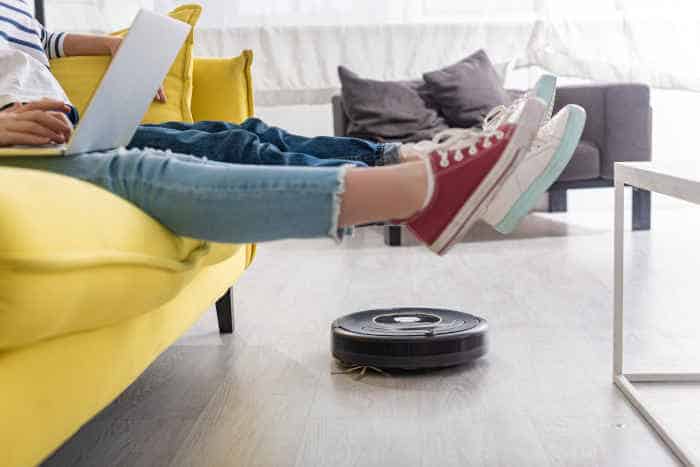 An iRobot vacuum cleaning under a couch while people relax on it.